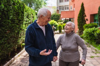 Mutlu yaşlı çift el ele tutuşuyor ve bir huzurevinin bahçesinde yürüyor güneşli bir günün tadını çıkarıyorlar.