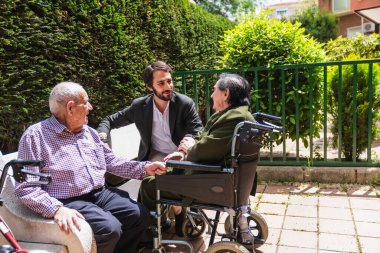 Doktor tekerlekli sandalyedeki yaşlı bir çiftle konuşuyor. Huzurlu bir huzurevinin bahçesinin keyfini çıkarıyor.