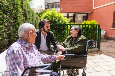 Doktor tekerlekli sandalyedeki mutlu yaşlı çiftin elini tutuyor, açık hava huzur evinde güneşli bir günün tadını çıkarıyor.