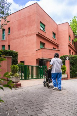 Bakıcı, yaşlı bir insanın tekerlekli sandalyesini açık mavi gökyüzünün altında pembe duvarlı ve yeşil aksanlı bir huzurevinin yakınına itiyor.
