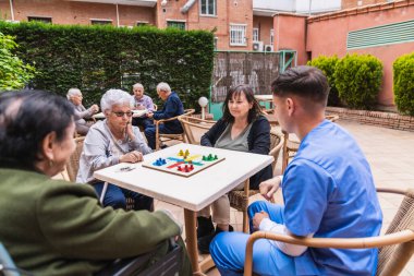 Hemşire, kız ve yaşlı kadınlar bir huzurevinin bahçesinde masa oyunu oynuyorlar, boş zamanların ve sosyal etkileşimin tadını çıkarıyorlar.