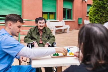 Tekerlekli sandalyedeki mutlu yaşlı kadın huzurevinin bahçesinde hemşirelerle masa oyunu oynuyor.