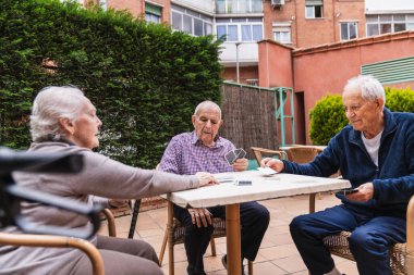Yaşlı bireyler dışarıda bir huzurevinde kart oynuyorlar, dingin bir ortamda sosyal etkileşimi ve eğlenceyi teşvik ediyorlar.
