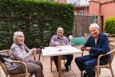 Üç kıdemli arkadaş dışarıda bir bakımevinin bahçesinde kart oynuyorlar. Sosyal etkileşimi ve aktif yaşlanmayı teşvik ediyorlar.