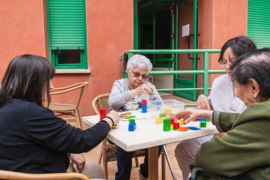 Güneşli bir günde, açık bir bakımevinde renkli bloklarla iş birliği yapan yaşlı kadınlar sosyal etkileşimi teşvik ediyorlar.