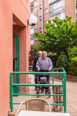 Hemşire, huzurevinin bahçesinde Walker 'la birlikte yaşlı bir adama yardım ediyor. Hareket kabiliyetini ve refahını güvenli bir ortamda teşvik ediyor.