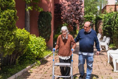 Erkek aile, güneşli bir günde bir huzurevinin bahçesinde yürüteç kullanan gülümseyen yaşlı kadına yardım ediyor.