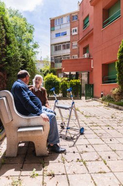 Aile, huzurevinin bahçesindeki bankta oturan yaşlı bir kadına bakımı ve desteği sağlar.