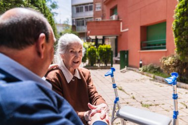 Aile ve kıdemli kadın, bir bakıcı ile açık hava ortamında sohbet ediyor, bakımevinde arkadaşlığı ve bakımı ön plana çıkarıyor.