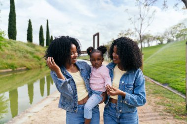Afrikalı ikiz kardeşler şehir parkında birlikte yürürken, güneşli bir günde doğa tarafından çevrelenmiş, yeğenlerine neşeyle sarılıyorlar.