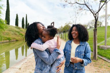 Afrikalı anne ve ikiz kız kardeşi parkta yürüyüp, küçük kızlarıyla gülüşüyor ve birbirlerine şefkat gösteriyorlar.
