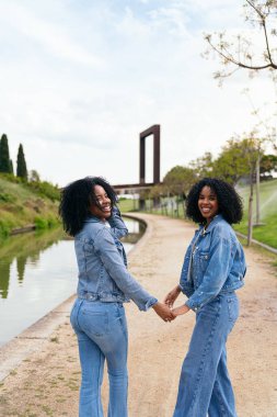 Afrikalı iki neşeli ikiz kardeş kot ceket ve kot pantolon giyip, el ele tutuşup, yeşilliklerle dolu güneşli bir parkta geziniyorlar.