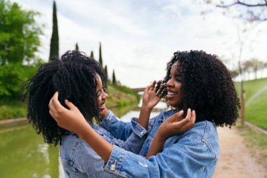 Afrikalı iki mutlu ikiz kardeş, kıvırcık saçlarına sarılıp dokunuyorlar. Parkta güneşli bir günün tadını çıkarıyorlar.