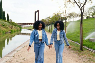 Kot ceket ve kot pantolon giyen, el ele tutuşan ve yeşil bir parkta kanal boyunca yürüyen mutlu ikiz kardeşler.