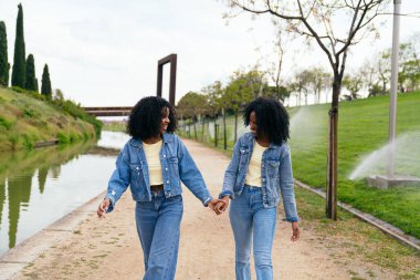 Sıradan kıyafetler giyen Afrikalı iki mutlu ikiz kardeş el ele tutuşup nehrin kenarındaki parkta yürüyorlar.