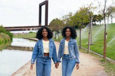 Kot ceket ve kot pantolon giyen, el ele tutuşan ve şehir parkında bir kanalın yanında yürüyen iki mutlu ikiz kardeş.