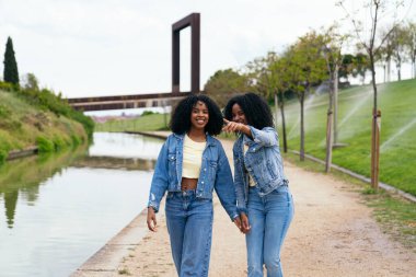 Kot ceket ve kot pantolon giyen, el ele tutuşan ve parkta yürürken kanalı gösteren iki mutlu ikiz kardeş.