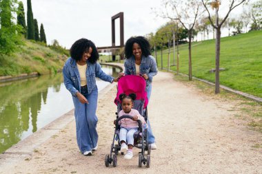 Parkta güneşli bir günün tadını çıkaran ikiz anneler, kanalda gezinirken, mutlu bir şekilde yeğenlerini bebek arabasına bindiriyorlar.