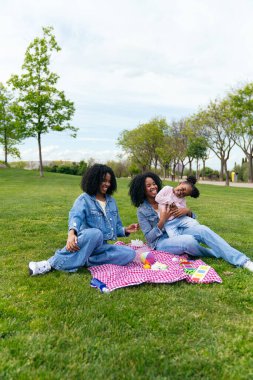 İki anne, güneşli bir günde kızlarıyla parkta pikniğin tadını çıkarıyor, birlikte oynuyor ve gülüyorlar.