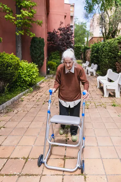 Yaşlı kadın huzurevinin bahçesinde yürüteç kullanıyor, temiz havanın ve güneşin tadını çıkarıyor.