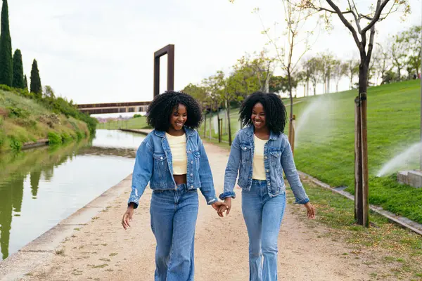 Kot ceket ve kot pantolon giyen, el ele tutuşan ve yeşil bir parkta kanal boyunca yürüyen mutlu ikiz kardeşler.