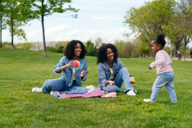 İkiz kardeşler parkta bir battaniyenin üzerinde oturuyor ve onlara doğru koşan yeğenleriyle oynuyorlar.