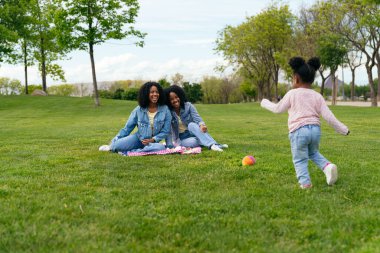 İki anne çimenlerde oturmuş, kızlarının neşeli, renkli bir baloya doğru koşuşunun keyfini çıkarıyorlar.
