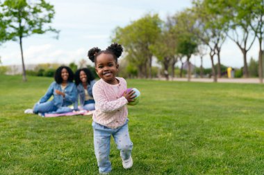 Mutlu bir çocuk, elinde renkli bir baloyla parkta çimenlerde koşarken aile üyeleri arka planda pikniğin tadını çıkarırken.