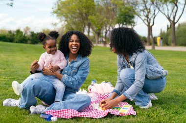Anne ve teyze birlikte gülüyor ve oynuyorlar. Canlı yeşil bir parkta yeğenleriyle güneşli bir pikniğin tadını çıkarıyorlar. Aile mutluluğunu kutluyorlar.