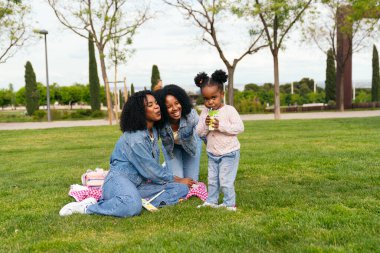 Afrikalı iki anne ve kızları parkta keyifli bir pikniğin tadını çıkarıyor, meyve suyunu yudumluyor ve sıcak güneşin altında kahkahalarını paylaşıyorlar.