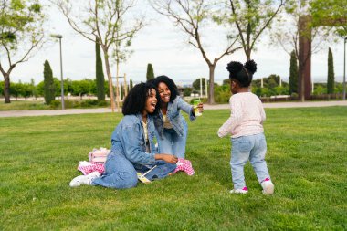 İki ikiz kardeş parkta piknik yaparken yeğenleriyle eğleniyorlar, güneşli bir günün tadını çıkarıyorlar.