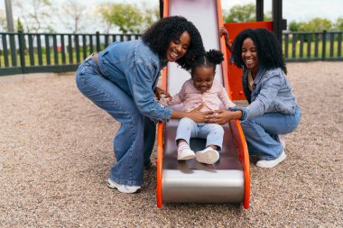 Afrika 'lı iki neşeli anne, kızlarına neşeyle oyun alanından kayarken yardım ediyor. Sevgi ve kahkaha anlarını paylaşıyorlar.