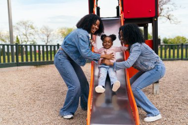 İki ikiz kız kardeş, güneşli bir günün tadını çıkarırken, yeğenlerinin oyun parkındaki kaydırağından kaymasına yardım ediyor.