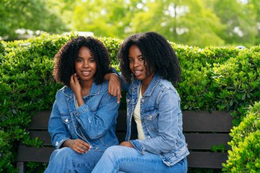 İki ikiz kardeş parkta bir bankta oturuyor, şık kot ceketler giyiyor ve neşeli gülümsemelerini paylaşıyor, açık havada güneşli bir günün tadını çıkarıyorlar.
