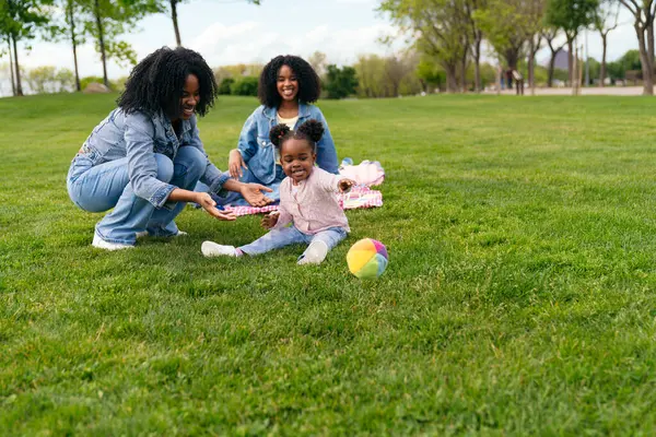 Afrikalı anne, teyze ve yeğen parkta piknik yapıyor, yeşil çimlerin üzerinde renkli bir topla oynuyorlar.