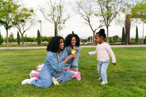 Afrikalı iki anne bir battaniyeye oturmuş kızlarına meyve suyu ikram ediyor yeşil bir parkta pikniğin tadını çıkarıyorlar.