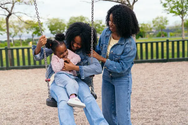 Anne ve teyze neşeli bir şekilde neşeli bir parkta salıncakta sallanan gülen bir kızı itiyor, aile eğlencesi ve birliktelik dolu güzel anlar yaratıyor.