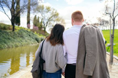 Parktaki gölette kol kola yürüyen genç bir çift güneşli bir bahar gününde romantik bir gezintinin tadını çıkarıyor.
