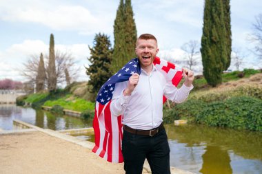 Beyaz tişörtlü, siyah pantolonlu, omuzlarının üzerinde büyük bir Amerikan bayrağı taşıyan, parkta nehir kenarında kutlama yapan kızıl saçlı bir adam.