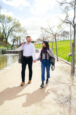 Genç çift el ele tutuşup parkta geziniyor güneşli bir günün sıcaklığında güneşleniyorlar ve birlikte vakit geçiriyorlar.