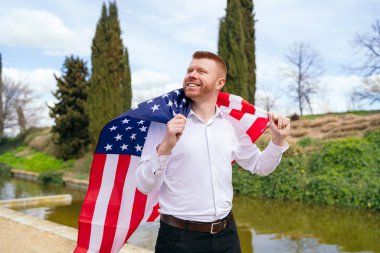 Beyaz tişörtlü ve siyah pantolonlu kızıl saçlı bir adam ulusal bayramı kutlamak için gölün kenarında Amerikan bayrağını omuzlarında tutuyordu.