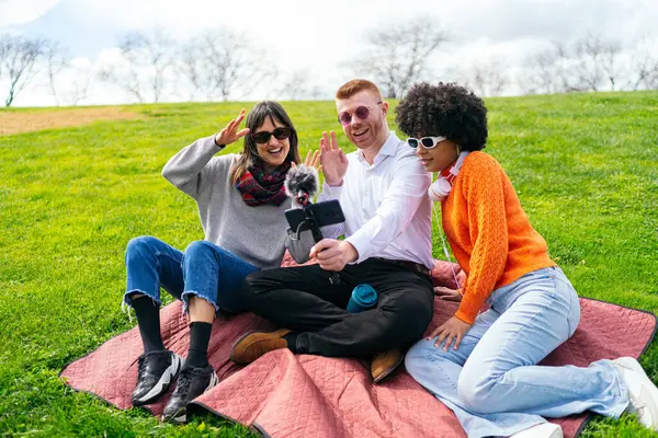 Akıllı telefon ve mikrofon kullanarak dışarıda bir video blogu kaydederken kameraya el sallayan üç içerik oluşturucu