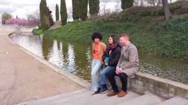 Üç farklı arkadaş, kanal, güneş ışığı ve sakin bir atmosfer yaratarak, yavaş çekim hareketi ve doğal kentsel arkaplan oluşturarak rahatça sohbet ediyorlar.