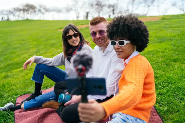 Üç genç içerik yaratıcısı, harici mikrofonu olan bir akıllı telefon kullanarak parkta bir video blogu kaydediyor.