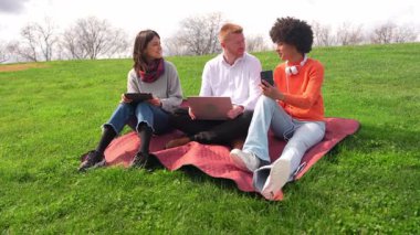 Üç üniversite öğrencisi ağır çekimde dizüstü bilgisayar, tablet ve akıllı telefon kullanıyor.