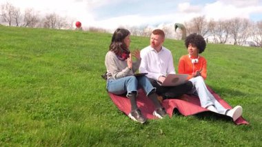 Öğrenciler açık havada bir proje üzerinde çalışıyorlar, dizüstü bilgisayar ve tablet kullanıyorlar, çimenli bir tepede bahar havasının tadını çıkarıyorlar.
