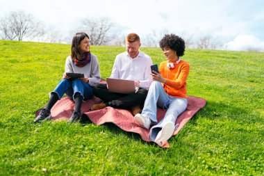 Üç öğrenci, çimenli bir parkta battaniyenin üzerinde rahatlarken elektronik aletler kullanıyorlar. Havanın tadını çıkarıyorlar.