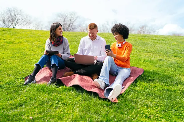 Üç öğrenci, çimenli bir parkta battaniyenin üzerinde rahatlarken elektronik aletler kullanıyorlar. Havanın tadını çıkarıyorlar.