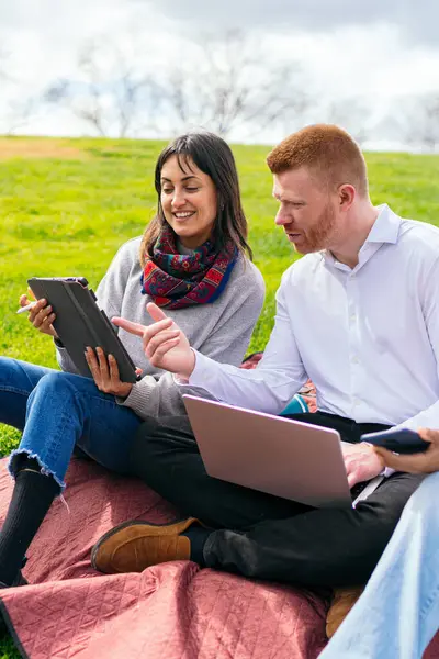 İki işadamı battaniyenin üzerinde oturuyor ve parkta dizüstü bilgisayar ve dijital tabletle uzaktan çalışıyor.