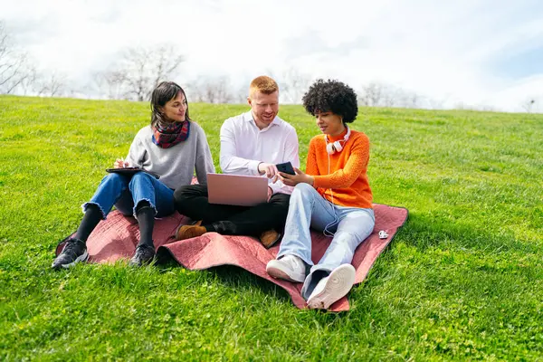 Üç öğrenci açık havada bir projede iş birliği yapıyorlar, bir dizüstü bilgisayar, tablet ve akıllı telefon kullanıyorlar, çalışırken temiz hava ve güneşin tadını çıkarıyorlar.